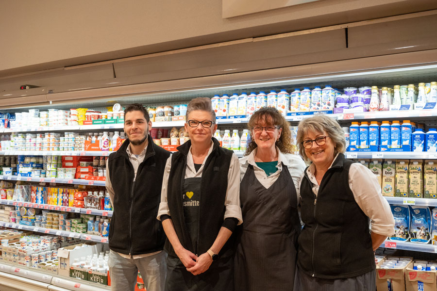 Vier Mitarbeitende stehen vor einem Kühlregal mit Milch- und Molkereiprodukten im Supermarkt.