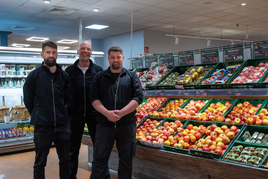 Drei Mitarbeiter stehen im Supermarkt vor der Obst- und Gemüseauslage mit Äpfeln und weiterem Obst.